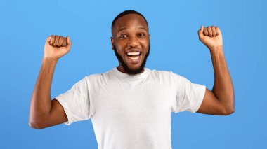 Zaferin sevinci. Neşeli Afro-Amerikan Yumruk Sallayan Adam Mavi Stüdyo Arkaplanındaki Kameraya Gülümseyen Adam. Siyahi Milenyum Adamı Başarı Kutlaması. Panorama