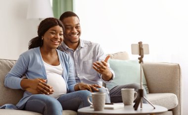 Neşeli siyah erkek ve kadın, blogcular, evden yayın yapan vlogcular, takipçilerle yaşam tarzlarını paylaşıyorlar, tripodlarda akıllı telefon kullanıyorlar, oturma odası içleri, hamilelik blogu konsepti