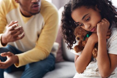 Kızgın siyah baba korkmuş küçük kızına bağırıyor oyuncağına sarılıyor ve aşağı bakıyor, iç mekan, yakın çekim. Aile içi kötü ilişkiler, Clildren kavramını kötüye kullanma.