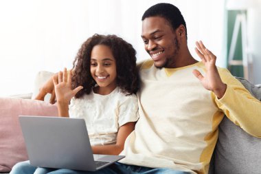 Mutlu siyah baba ve kız, evde birlikte vakit geçirirken aileleriyle video konferansı yapıyor, kanepede oturuyor ve dizüstü bilgisayar ekranına el sallıyor, iç mekan, fotokopi odası.