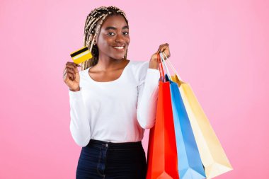 Sınırsız alışveriş konsepti. Renkli alışveriş torbaları taşıyan neşeli siyah kadın portresi ve kredi kartını parlak pembe stüdyo arka planında gösterme, kopyalama alanı