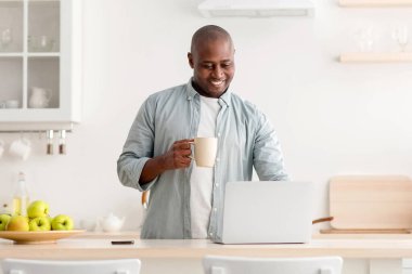 Kendini izole eden modern bir işçi. Kahvesini yudumlayan ve mutfaktaki masada dizüstü bilgisayarda çalışan, fincanlı Afro-Amerikalı mutlu bir adam.
