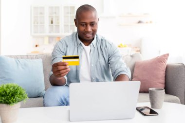 Kolay ödeme ve nakit ödeme konsepti. Kredi kartı tutan ve dizüstü bilgisayar kullanan mutlu Afro-Amerikalı adam, evdeki koltukta oturuyor, klavyeye, panorama, kopyalama alanına yazı yazıyor.