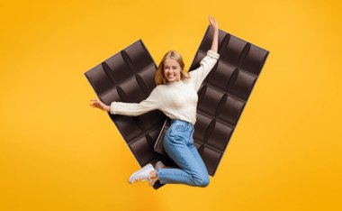 Genç bir kadın, büyük çikolatalarla çevrili canlı sarı bir arka plana heyecanla atlıyor. Neşeli duruşu bir neşe ve enerji hissini vurguluyor..