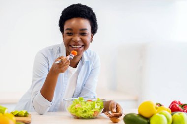 Modern mutfaktaki kameraya gülümseyen ve akşam yemeği için taze sebze yiyen mutlu siyah kadın. Beslenme, Sağlıklı Yemek ve Kilo Verme Tarifi