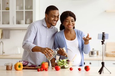 Mutlu siyahlar, evde sağlıklı yemek pişirirken ve cep telefonunu tripodda kullanırken aile yayını bekliyor. Neşeli Afrikalı Amerikalı hamile çift internetten aşçılık dersi alıyor.