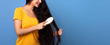 Gülümseyen genç Hintli kadının portresi uzun kıvırcık ve dalgalı saçlarına bakıyor, ahşap fırçayla tarıyor, mor stüdyo arka planında tek başına duruyor, bedava fotokopi alanı