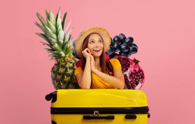 Hasır şapka ve parlak sarı gömlek giyen neşeli bir gezgin sarı bir bavulun üzerinde oturuyor..