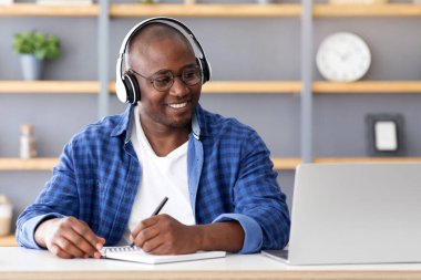 E-eğitim konsepti. Mutlu siyah olgun bir adam dizüstü bilgisayar kullanıyor ve notlar alıyor, kablosuz kulaklık kursları dinliyor ve ekranlara bakıyor, iç mekandaki masada oturuyor.