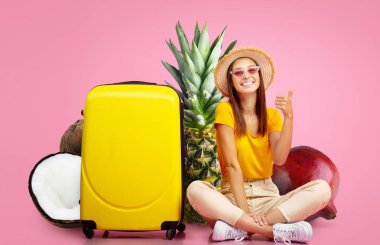 Genç bir kadın, parlak sarı bir bavulun yanında baş parmağıyla oturuyor. Arkasında ananas ve hindistan cevizi de dahil olmak üzere tropikal meyveler var. Hepsi canlı pembe arka planda..