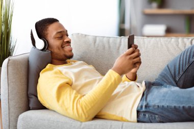 Neşeli Afro-Amerikalı adam evdeki koltukta dinlenirken cep telefonuyla video seyrediyor, kablosuz kulaklık kullanıyor, fotokopi çekiyor. Akıllı telefondan film izleyen neşeli siyahi adam.