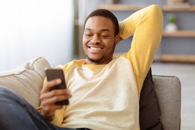 Evdeki kanepede uzanan, akıllı telefon kullanan, arkadaşlarıyla sohbet eden ya da internette oynayan, fotokopi çeken siyahi adamın yakın çekimi. Mutlu Afro-Amerikan genç adam internet sörfü yapıyor veya cep telefonunda sosyal medya kullanıyor.