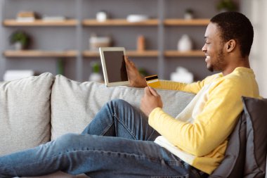 Kredi kartı ve dijital tabletli neşeli Afro-Amerikalı genç adam evdeki kanepeye oturmuş, fotokopi çekiyor. İnternetten alışveriş yapan mutlu siyah adam, ped, e-ticaret, maket