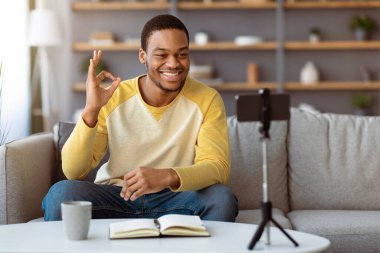 Neşeli genç siyahi adam evde otururken online ders alıyor, oturma odasında koltukta oturuyor ve cep telefonunu tripodun üzerinde kullanıyor, akıllı telefon kamerasına iyi bir hareket gösteriyor ve gülümsüyor, uzayı kopyalıyor.