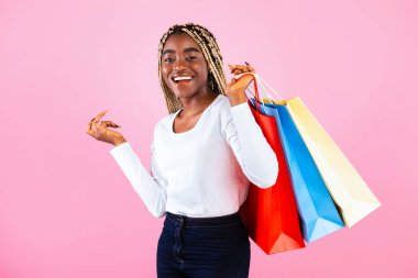 Alımlarını gösteren, elinde pembe stüdyo arka planında renkli alışveriş torbaları tutan güzel siyah bir kadın.
