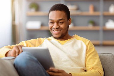 Oturma odasındaki kanepede oturan gülümseyen siyah adam dijital tabletin en yeni uygulamasını kullanarak, uzayı kopyaladı. Mutlu Afro-Amerikan erkeği evde eğleniyor, sosyal medyayı kontrol ediyor ya da arkadaşlarıyla sohbet ediyor.