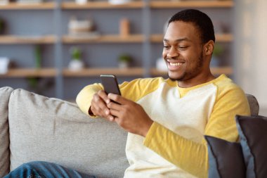 Evdeki kanepede oturan, akıllı telefon kullanan, arkadaşlarıyla sohbet eden, internette oynayan, kopya alan gülümseyen siyah adam. Mutlu Afro-Amerikan genç adam internet sörfü yapıyor veya cep telefonunda sosyal medya kullanıyor.
