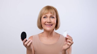 Hafif stüdyo arka planında pamuk ped ve ayna kullanarak yumuşak cilt temizleme yüzlü mutlu yaşlı bayan portresi tonik veya mikiler suyu ile. Tatlı olgun kadın kendine dikkat ediyor.