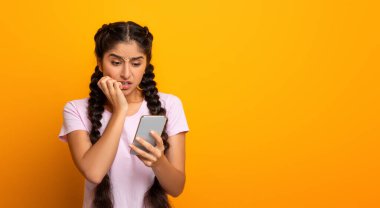 Haberler kötü. Endişeli Hintli genç bayan cep telefonuna bakıyor ve olumsuz yorumlar yapıyor. Sarı stüdyo arka planında, afişte, bedava fotokopi alanında tırnak yiyen kadın.