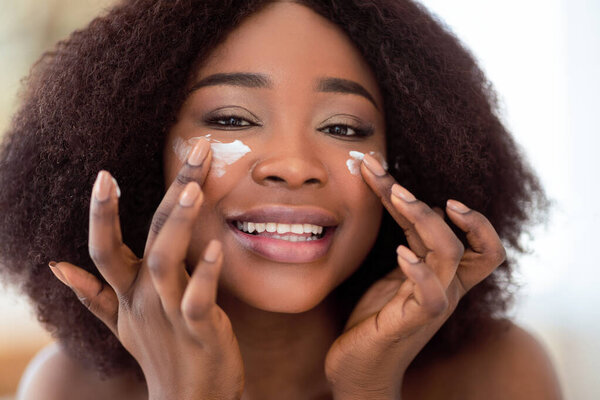 Portrait of lovely black woman applying face cream onto her cheeks, smiling at camera, indoors. Beautiful African American lady using moisturizing lotion, pampering her silky skin at home