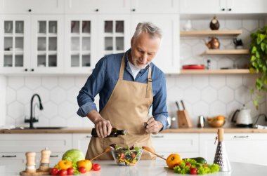 Gülümseyen kıdemli adam mutfakta taze sebze salatası pişiriyor, zeytinyağı ve baharat katıyor, önlük giymiş yaşlı beyefendi evde sağlıklı vejetaryen yemekleri yapıyor, uzayı kopyalıyor.