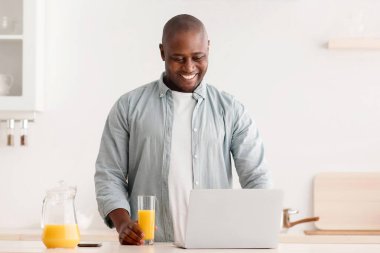 Olgun siyahi bir adam internette haber okuyor ve mutfakta portakal suyu içiyor. Hafta sonları sabah keyfine bakıyor ve evde dinleniyor. Boş bir alanda.