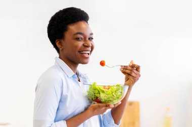 Evdeki modern mutfaktaki kameraya gülümseyen, dizüstü bilgisayar yiyen, salata yiyen siyah kadın. Gıda Bloğu ve Kilo Verme Sitesi. Sağlıklı Beslenme ve Diyet Tarifleri İnternet 'te. Seçici Odaklanma