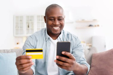 Çevrimiçi alışveriş. Kredi kartı ve akıllı telefonlu neşeli Afro-Amerikalı adam evdeki kanepede oturuyor, internetten alışveriş yapıyor, yakın plan çekiyor. E-ticaret kavramı