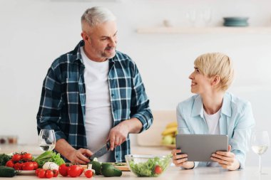Birlikte mutfakta yemek pişirmek, internette yemek tarifi aramak, online blog izlemek. Mutlu orta yaşlı adam salata için sebze keser, tabletli kadın mutfak içi yemek dersi, boş alan.