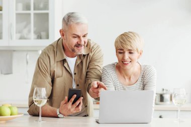 Video görüşmesi, online alışveriş, evdeki cihazlarla çalışma. Telefonu olan mutlu yaşlı bir adam, minimalist mutfakta, içi meyve kadehleriyle dolu, boş bir yerde, olgun bir bayana parmak dizüstü bilgisayarı gösteriyor.