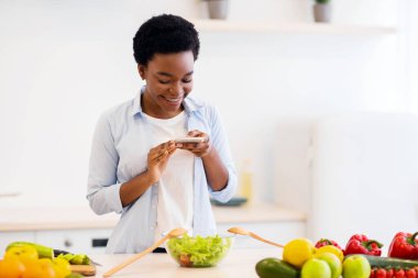 Yemek blogu. Siyahi kadın blogcu telefondaki sağlıklı yemeğin fotoğrafını çekiyor modern mutfakta duran salata pişiriyor. Beslenme ve Kilo Blog Konsepti. Diyet ve Zayıflama Uygulaması