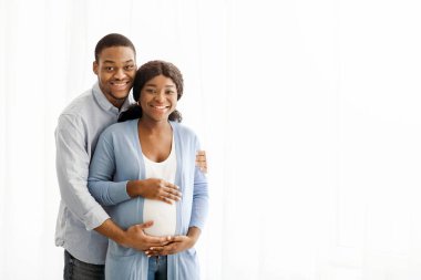 Mutlu siyah çift bebek bekliyor, beyaz arka planda poz veriyorlar, kameraya sarılıyorlar ve gülümsüyorlar, fotokopi alanı olan bir panorama. Pozitif Afro-Amerikan hamile ailesi evde sarılıyor.