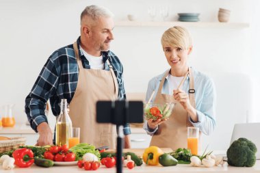 Çift birlikte yemek pişiriyor, yemek tarifi veriyor, video bloğu ve sosyal medya videolarını akıllı telefon kamerası ve tripodla kaydediyor. Mutlu bir eş ve koca mutfak içinde yemek hazırlar, boş bir alan, bulanık.