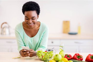 Diyet Tarifleri Mobil Uygulaması. Modern mutfaktaki yemek hazırlama uygulaması ile akıllı telefon kullanan mutlu Afrikalı kadın. Beslenme ve Teknoloji Konsepti