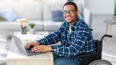 Tekerlekli sandalyedeki neşeli siyahi adam internetteki iş için bilgisayarını kullanıyor ve internet üzerinden iş görüşmesi yapıyor. Uzak bir projede çalışan özürlü bir Afrikalı Amerikalı.