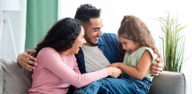 Üç kişilik neşeli bir Arap ailesi evde eğleniyor. Orta Doğulu ebeveynler küçük kızlarını gıdıklıyor ve oturma odasında dinlenirken birlikte gülüyorlar, web pankartı.