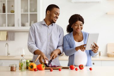 Smiling black pregnant couple using digital tablet while cooking healthy food together in kitchen at home, copy space. Cheerful african american expecting family reading recipes online, using pad