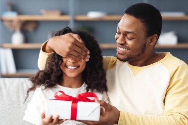Ev kavramında doğum günü kutlaması. Sevgi dolu siyah baba kızına hediye veriyor, avuçlarıyla gözlerini kapatıyor, oturma odası içi, fotokopi odası. Mutlu Afro-Amerikan ailesi doğum gününü kutluyor.
