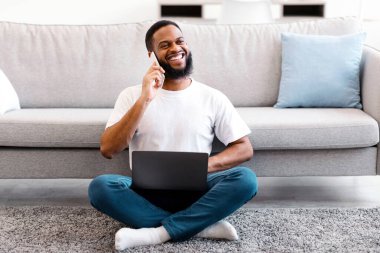 Cep telefonuyla konuşan Afrikalı Amerikalı adam dizüstü bilgisayarla oturmuş evden uzaktan çalışıyor. Modern İletişim ve Serbest Yaşam Tarzı. Uzak Çalışma