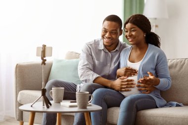 Gülümseyen Afro-Amerikan genç adam ve hamile kadın evden arkadaşlarıyla veya akrabalarıyla görüntülü görüşme yapıyorlar, iyi haberleri paylaşıyorlar, kanepede oturuyorlar, cep telefonlarını tripoda kullanıyorlar,