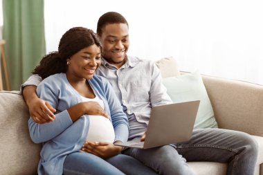 Hamile siyah çifti sevmek, dizüstü bilgisayar kullanmak, evde film izlemek, oturma odasında koltukta oturmak, Afrikalı Amerikalı bir kadının kocasıyla vakit geçirmesini beklemek,