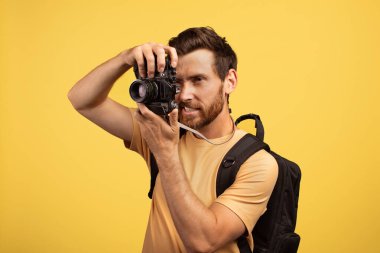 40 'lı yaşlarda sıradan bir orta yaşlı adamın elinde özel bir anı yakalamaya odaklanmış bir kamera var. Basit bir tişört giyiyor ve sırtında parlak sarı bir arka planda duran bir sırt çantası var..