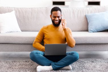 Evdeki oturma odasında dizüstü bilgisayarın üzerinde çalışırken cep telefonuyla konuşan Afrikalı adam. Her zaman temastayız. Serbest Yaşam Tarzı ve Uzaktan Çalışma