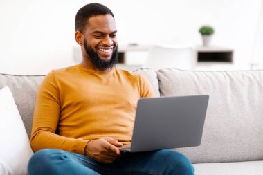 Siyah adam dizüstü bilgisayar kullanıyor koltukta uzaktan çalışıyor ve evdeki interneti tarıyor. Modern Teknoloji, Serbest Yaşam Tarzı ve Uzaktan Çalışma Çevrimiçi Kavramı.