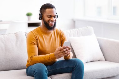 Kulaklık takan, akıllı telefon kullanan, internetten müzik dinleyen ya da evdeki koltukta oturan siyah adam. Telefon Kullanıcısı Kablosuz Kulaklıklardaki Sesin Tadını Çıkartıyor