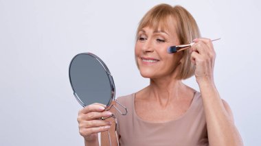 Yüzüne dekoratif makyaj uygulamak için kozmetik fırça kullanan hoş bir bayan stüdyo arkaplanındaki aynaya bakıyor. Olgun bir kadın kızarıyor, kendine gülümsüyor.