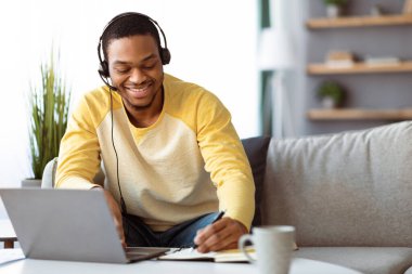 Evdeki çevrimiçi eğitime katılan pozitif siyah bir adam, sehpanın önünde dizüstü bilgisayarla oturuyor, kulaklık kullanıyor, notlar alıyor, kopyalama alanı kullanıyor. E-eğitim, çevrimiçi sınıf, eğitim kavramı