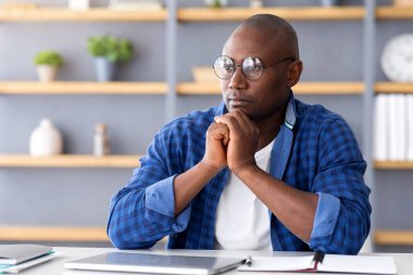 İş planını düşünüyorum. Düşünceli siyahi girişimci, yeni bir proje üzerinde çalışıyor. İş yerinde, düşünceli bir yüz ifadesiyle oturuyor, yan gözle bakıyor.