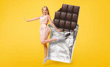 Neşeli genç bir kadın, parlak bir folyonun yanında dururken gülümseyen büyük bir çikolata çubuğuyla poz veriyor. Parlak sarı arka plan neşeli atmosferi güçlendirir..