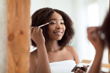 Şehvetli genç siyahi kadın evde aynanın yanında aplikatörle göz farı sürüyor. Dekoratif makyaj yapan güzel Afrikalı Amerikalı kadın, organik kozmetik ürünler kullanıyor.